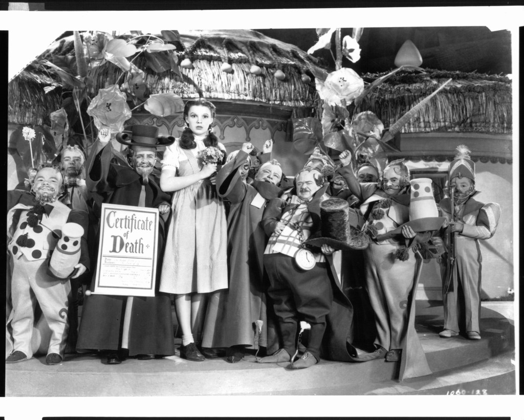 Meinhart Raabe holding a death certificate and Judy Garland surrounded by munchkins in a scene from the film 'The Wizard Of Oz', 1939. (Wizard of Oz behind the scenes)