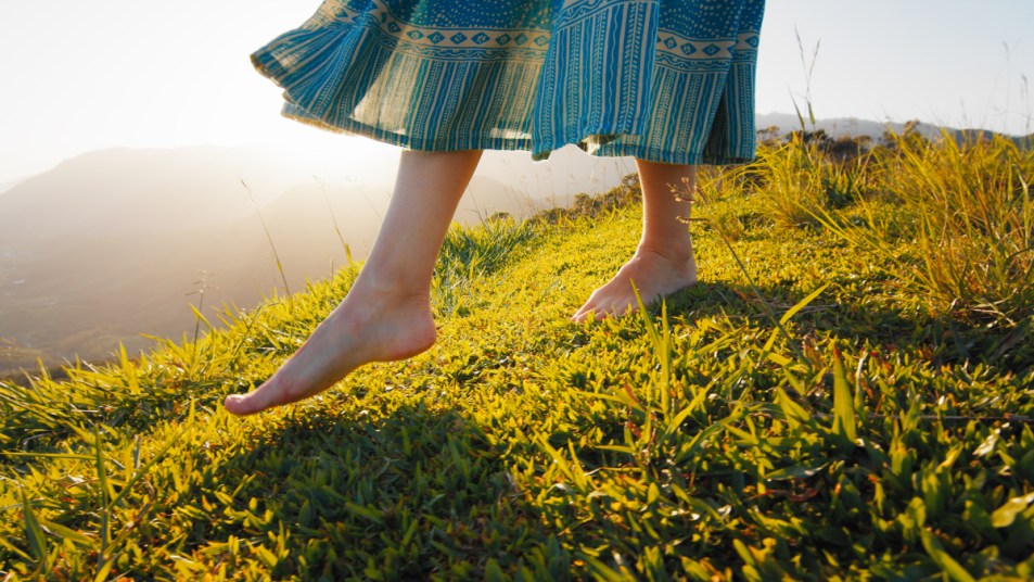 A close-up of a woman in a flowing dress walking barefoot in the grass to enjoy the health benefits