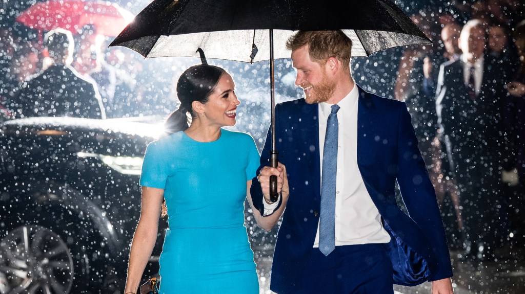 Prince Harry and Meghan smiling at each other under and umbrella