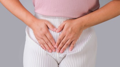 A close up of a woman in a pink top and white pants, holding her hands on her uterus before trying bacterial vaginosis home remedies