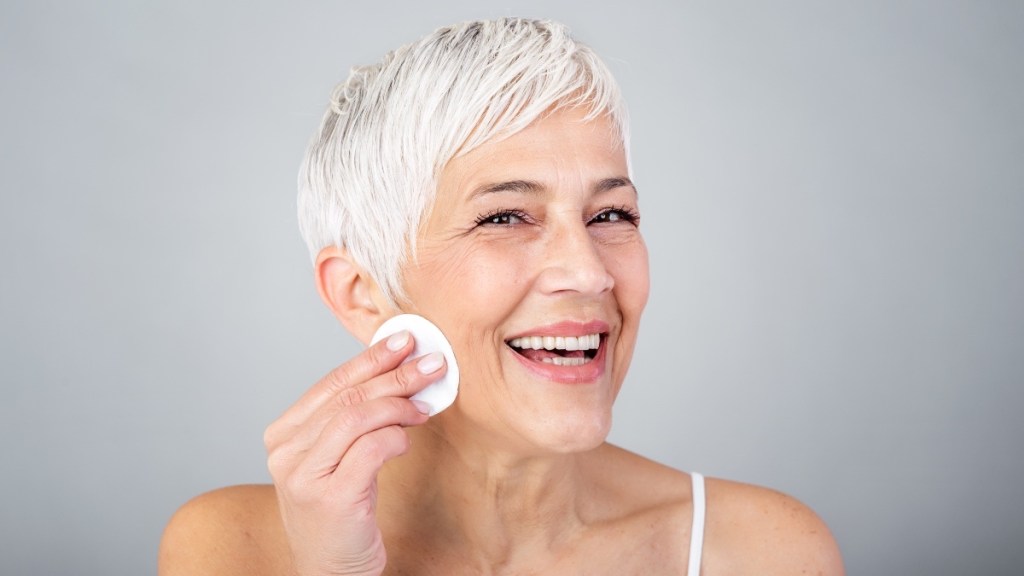 A woman with short grey hair swiping a cotton round on her face to apply apple cider vinegar