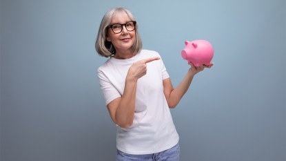 mature woman in tee shirt and jeans pointing at a piggy bank