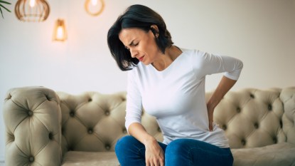A woman with dark hair and a white top sitting on a couch while holding her lower back, which is in pain and can be helped with self-care