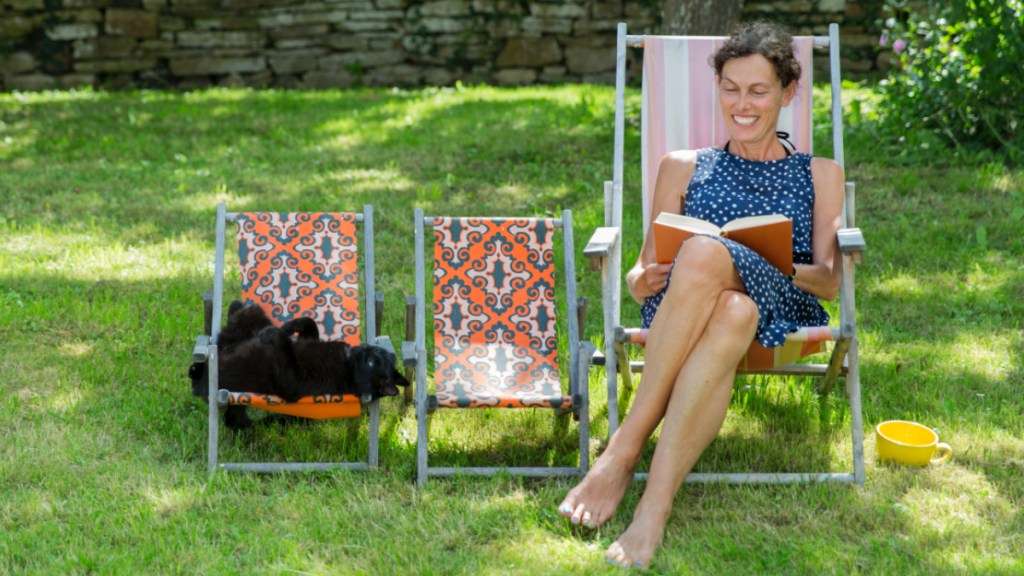 A barefoot woman reading a book in a chair beside a cat in a chair in the grass