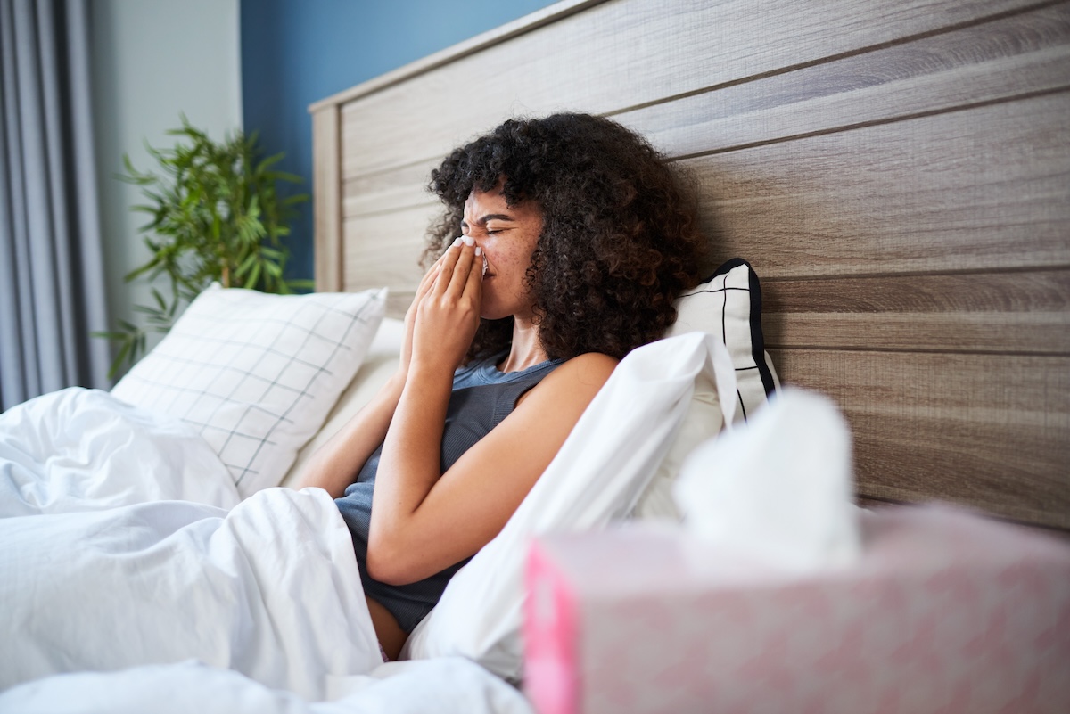 A person rests in a bed in a well-lit bedroom. They are using a tissue, indicating symptoms such as a cold or allergy. The environment suggests comfort and recovery in a modern home.