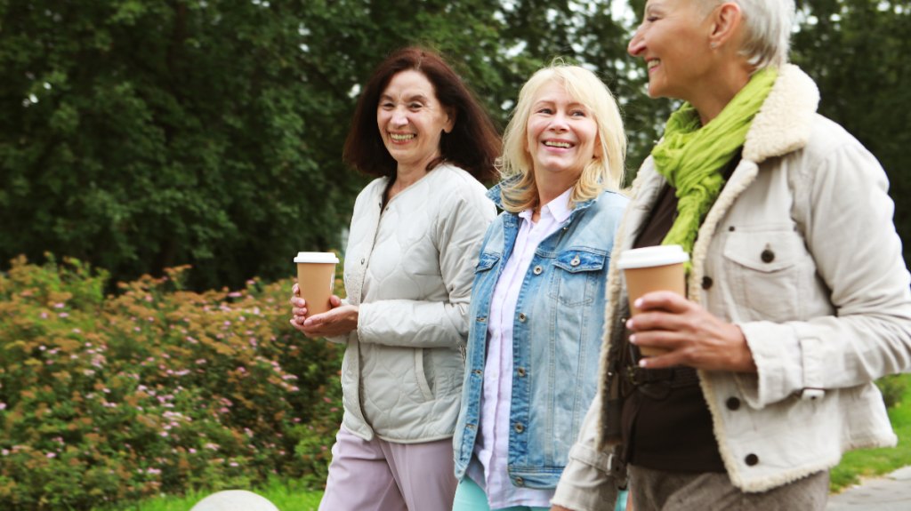 three middle-aged women are happily walking oudoors, coffees in hand