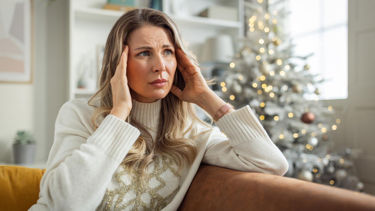 stressed-out woman massages her temples as she sits with a Christmas tree in the background