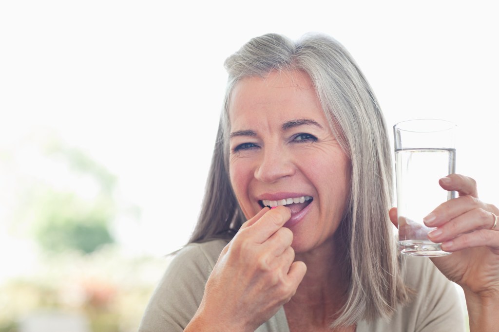 older woman smiles as she prepares to take a vitamin while holding a glass of water