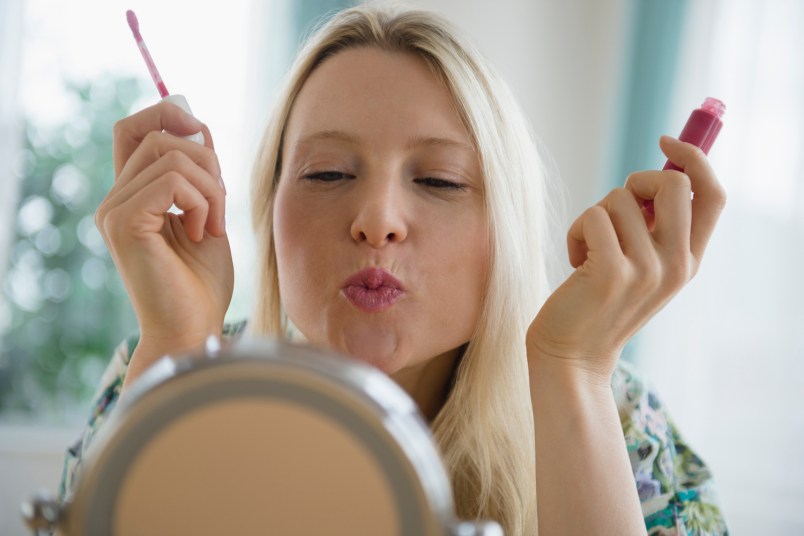 woman holding lip gloss and puckering her lips at a mirror