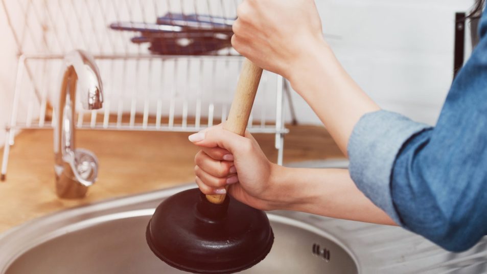 Cropped view of woman in denim shirt using plunger in sink