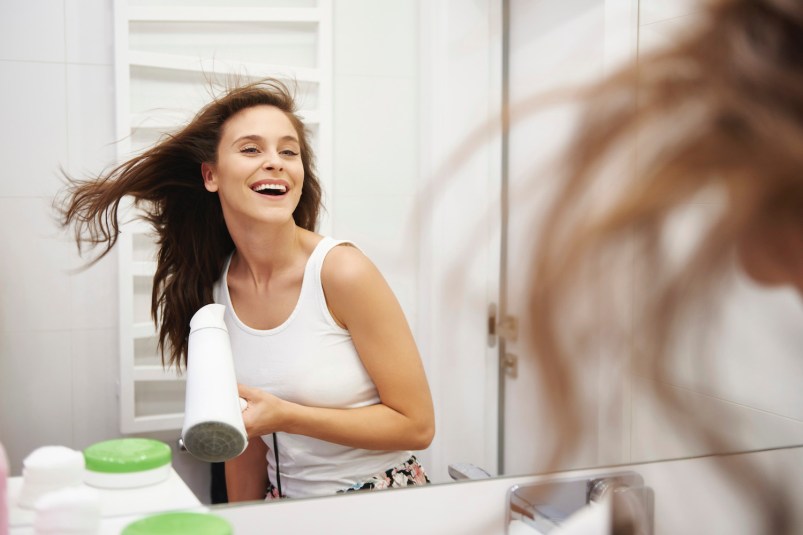 Mirror image of laughing woman blow-drying her hair in the bathroom