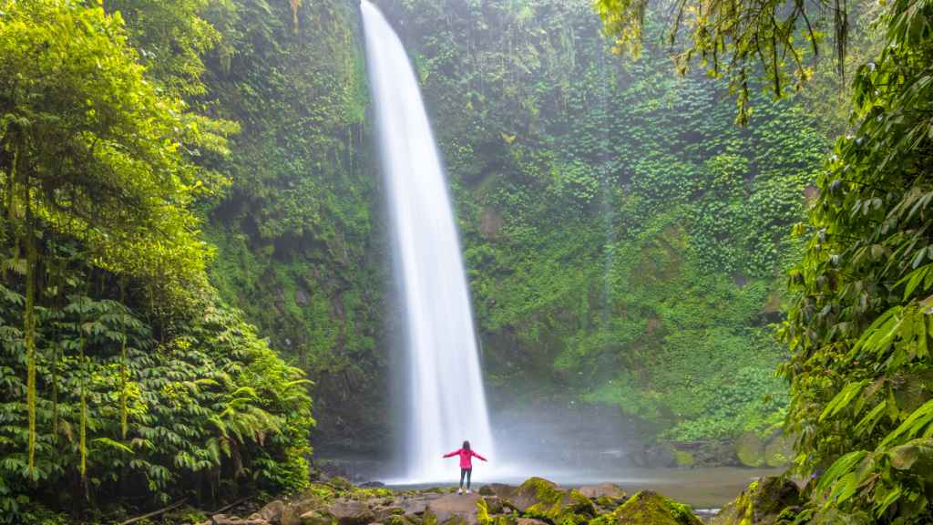 woman stands dwarfed at the bottom of a majestic waterfall, as she happily raises her arms and lets go of regrets