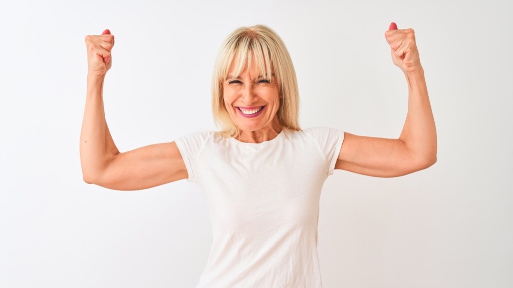 Blonde woman over 50  who supplements with vitamin D, in white t-shirt flexing her bicep muscles