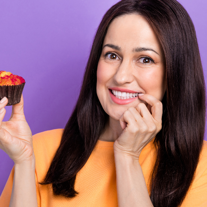 middle-aged woman holds a cupcake in one hand while pressing her fingers playfully to her lips with the others, as if debating whether to eat the cupcake