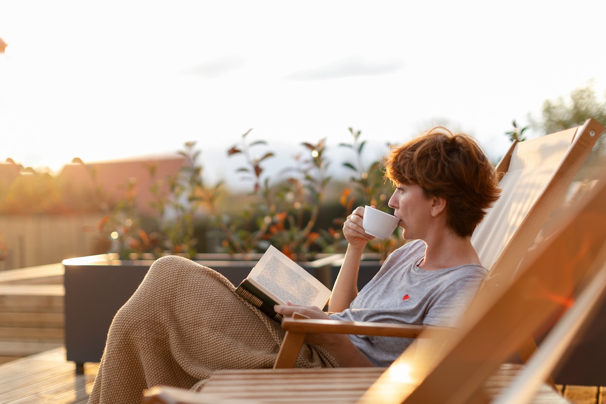 Beautiful woman is sitting on patio on the deck chair and enjoying beautiful surrounding while drinking coffee or tea and reading a book