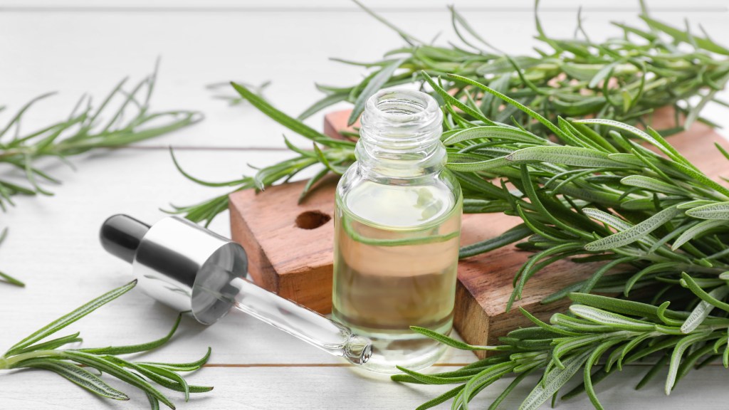 Fresh green rosemary and bottle of essential oil on white wooden table next to wood cutting board