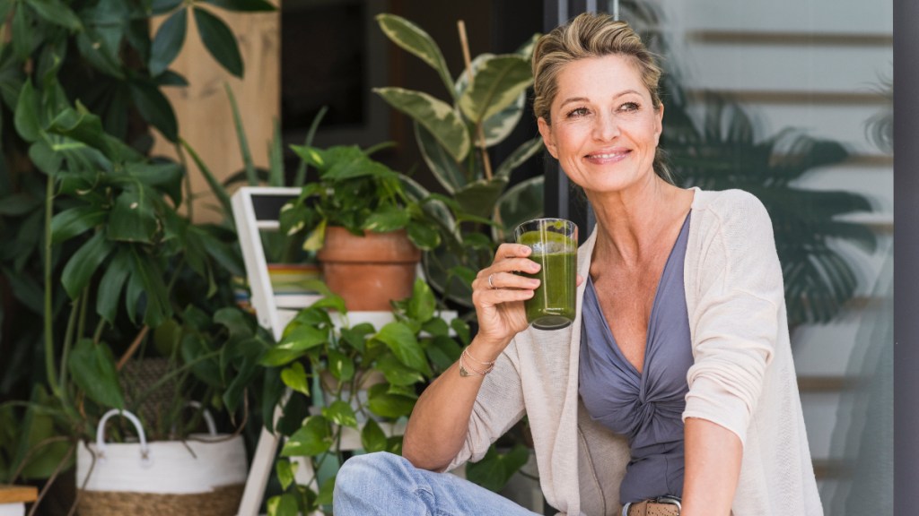 A blonde woman holding a green smoothie while sitting in front of green plants