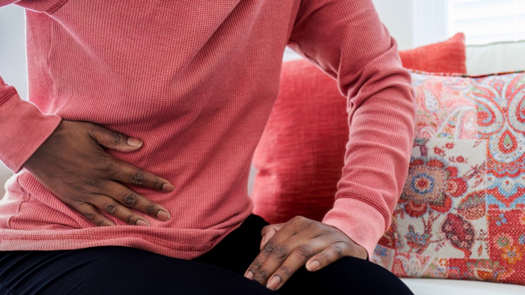 A close-up of a woman in a pink top and black pants holding her hand to her stomach in pain