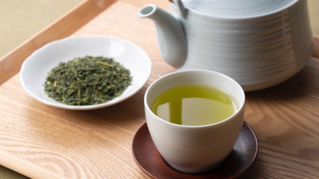 A cup of green tea next to a teapot and bowl of loose green tea leaves