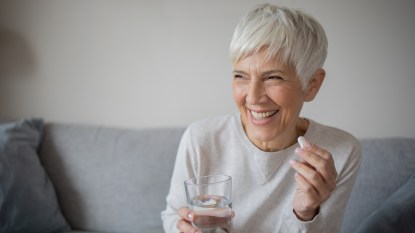 A smiling, grey-haired woman holding the best vitamin for anxiety and a glass of water while sitting on a couch