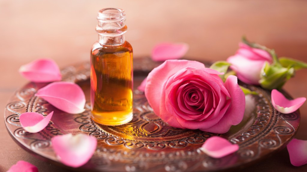 A vial of rose essential oil on a tray next to a pink rose and rose petals