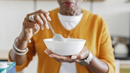 Close up shot of a senior African American woman holding a bowl of cereal for breakfast while standing in the kitchen. Selective focus on the bowl and spoon in her hands.