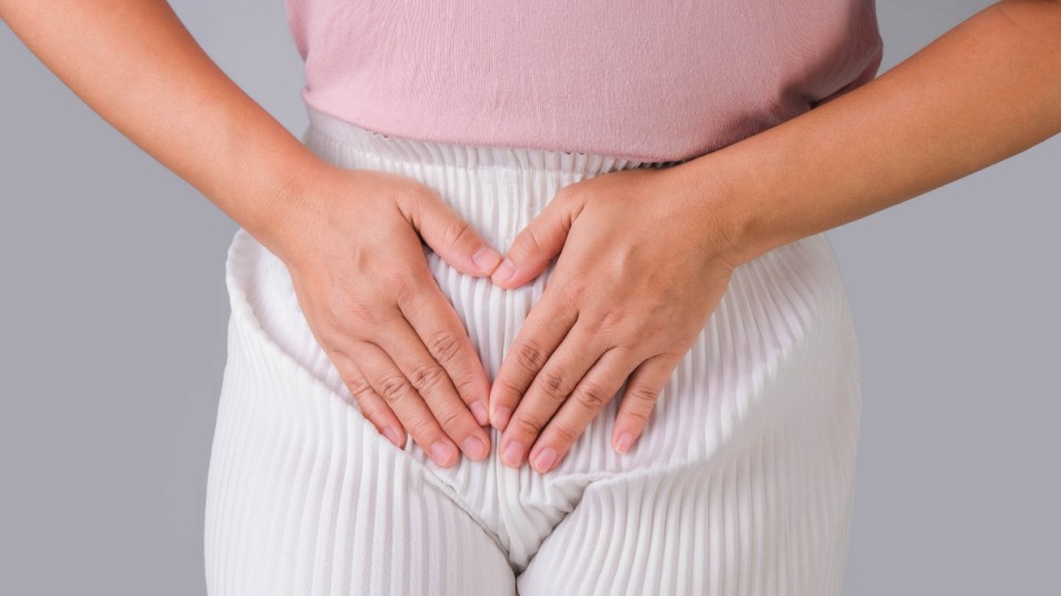 A close up of a woman in a pink top and white pants, holding her hands on her uterus before trying bacterial vaginosis home remedies
