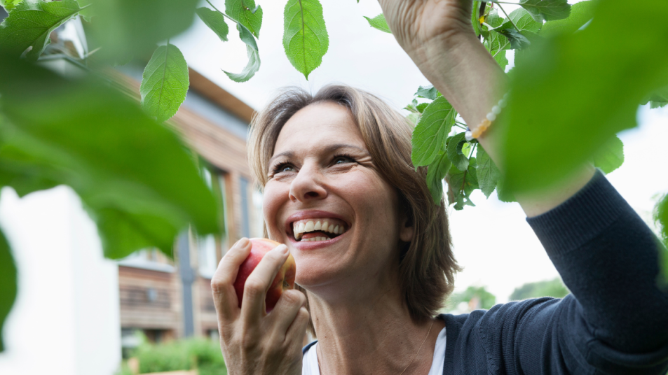 smiling woman eats an apple full of fiber, standing outside by the green leaves of a tree
