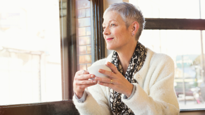 mature woman with short hair gazing out window and smiling, holding a cup of coffee