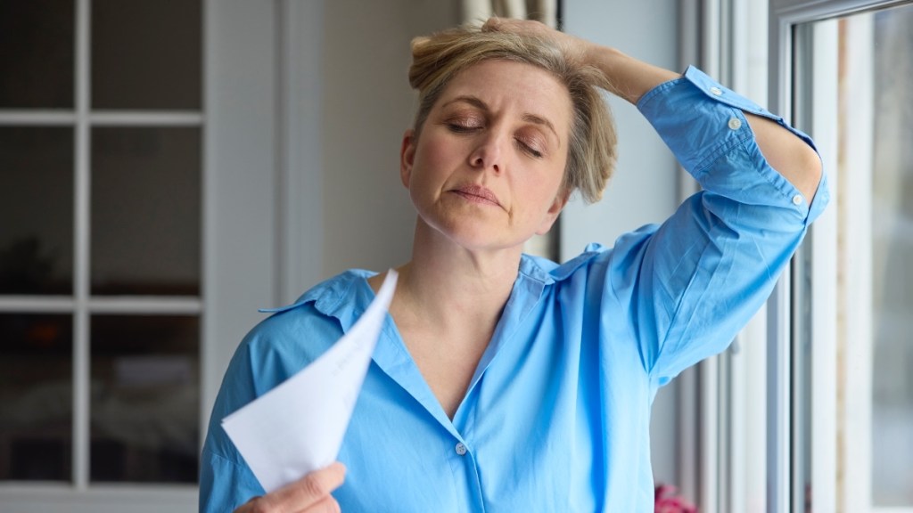 A blonde woman in a blue shirt holding her hair up and fanning herself with paper due to a hot flash