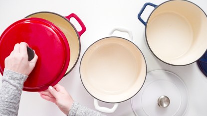 Hands removing lid from red dutch oven next to white and blue dutch ovens