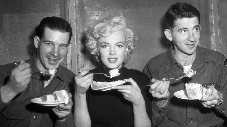 American actress Marilyn Monroe (1926 - 1962) tries some cake in the Enlisted Men's Mess Hall at Headquarters Company, 2nd Infantry Division, near Seoul, South Korea, during her wartime entertainment tour of Korea, 18th February 1954. With her (right) is Sgt. Harold Crawford of Columbus, Ohio.