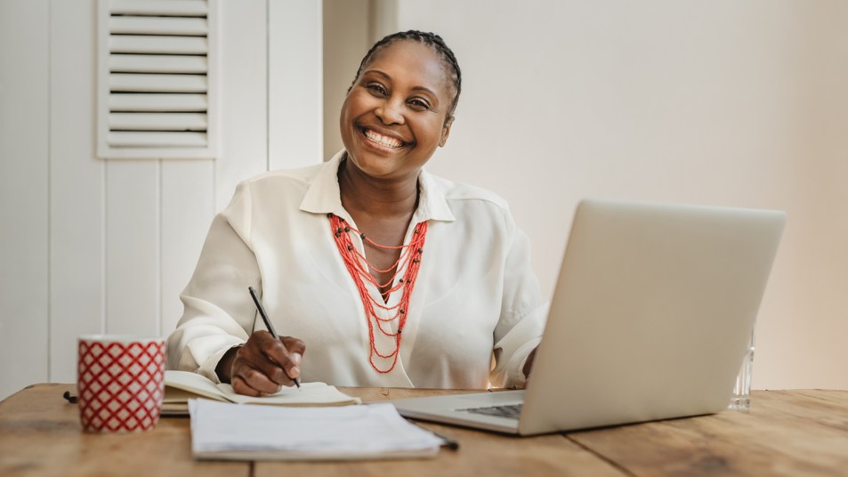 Smiling Black woman working on a laptop and writing notes while sitting at a desk in her home office