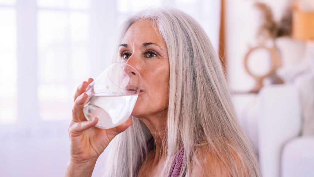 A woman with long, grey hair drinking a glass of water to help stop a runny nose in 5 minutes