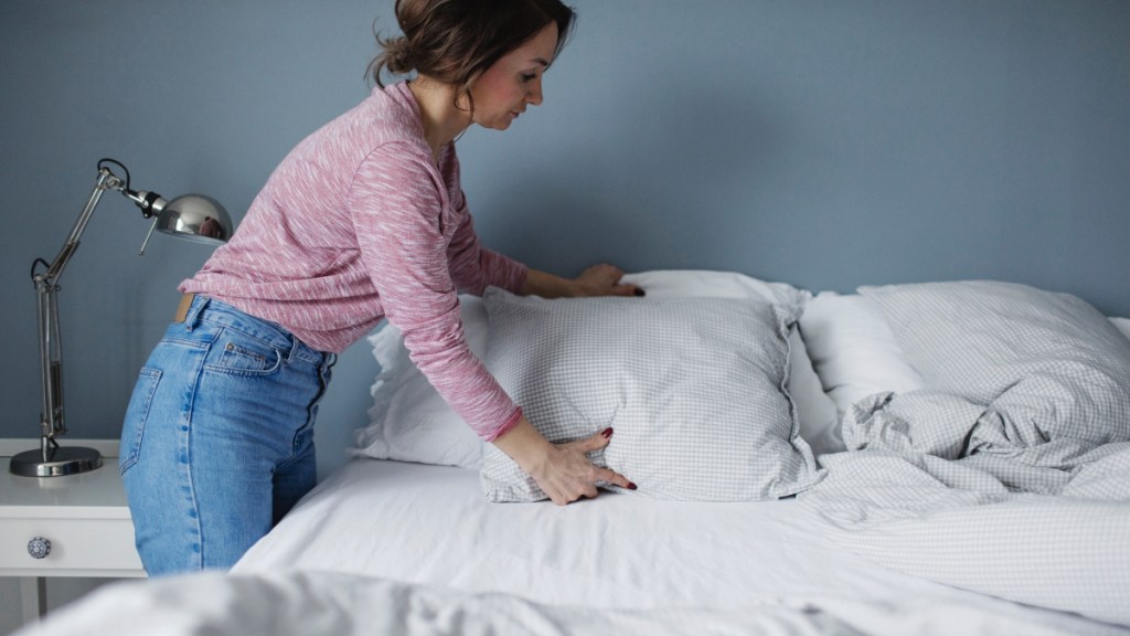 A woman in a sweater and jeans fluffing a pillow on her bed to help stop a runny nose in 5 minutes