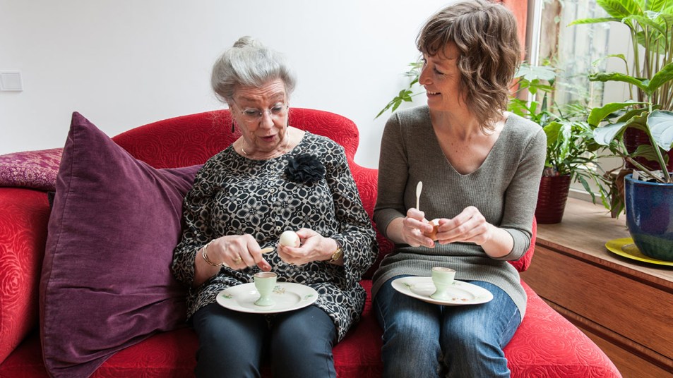 One elderly white woman with white hair enjoys a soft boiled egg with a younger white woman who sits next to her, also eating an egg.