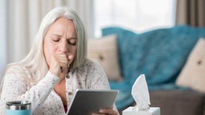 senior woman with straight gray hair coughing into her hand, holding a tablet, with tissues beside her