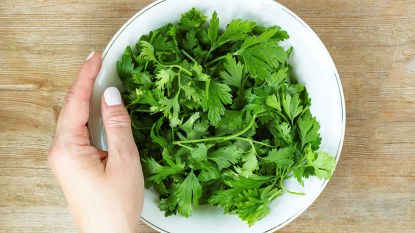 White female hand shown holding a bowl of cilantro against a wooden background.