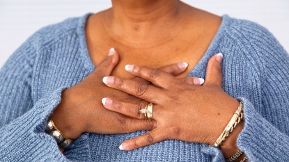 Mature black woman in periwinkle blue sweater folds hands over her heart.