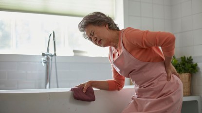 Mature asian woman holds her lower back in pain while cleaning her kitchen sink.
