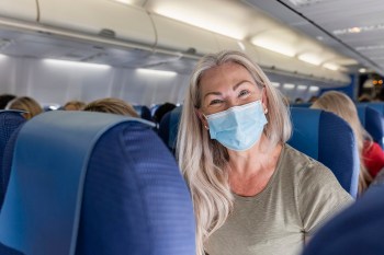 Mature caucasian woman sits on a plane wearing a mask and smiling excitedly beneath it.