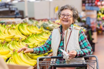 Mature asian woman smiles near a display of fresh bananas while grocery shopping.
