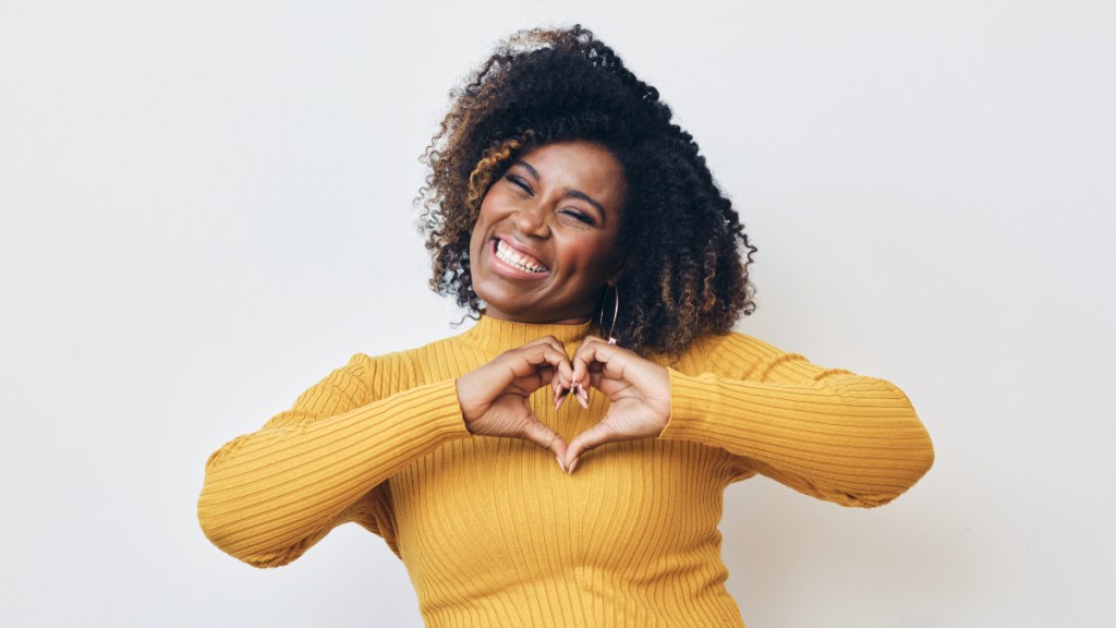 An African American woman in a gold shirt making a heart with her hands in front of her chest