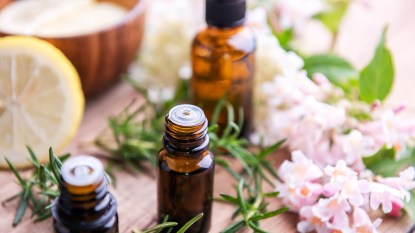 Clear, brown bottles of essential oil sit on a surface surrounded by fresh herbs and flowers, a lemon slice, and wooden bowl in the background.