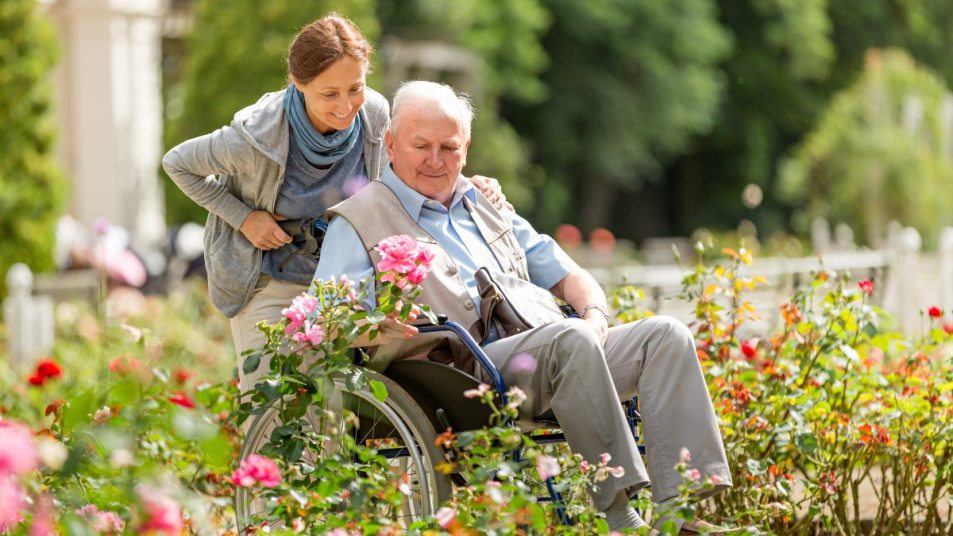 mature woman pushing wheelchair of a loved one in a nursing home, outside in flower garden