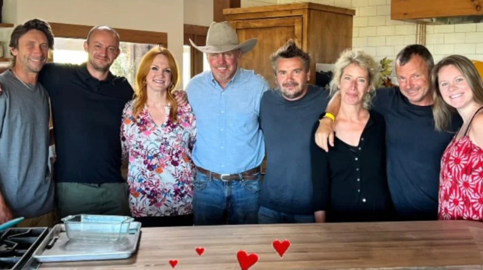 Seven-member Pioneer Woman cooking show cast pose hugging behind kitchen counter on Ree Drummond TV set