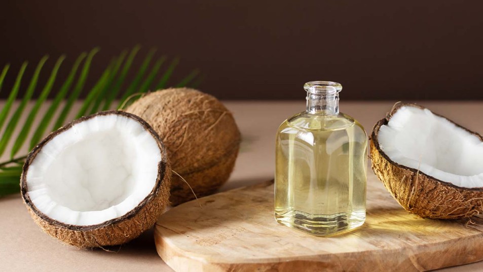 Bottle-of-coconut-oil-and-fresh-coconuts-with-palm-leaf-on-wooden-board-over-brown-background