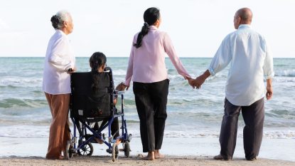 back view of a senior woman in a wheel chair and three family members around her, on the beach, experiencing the joys of travel