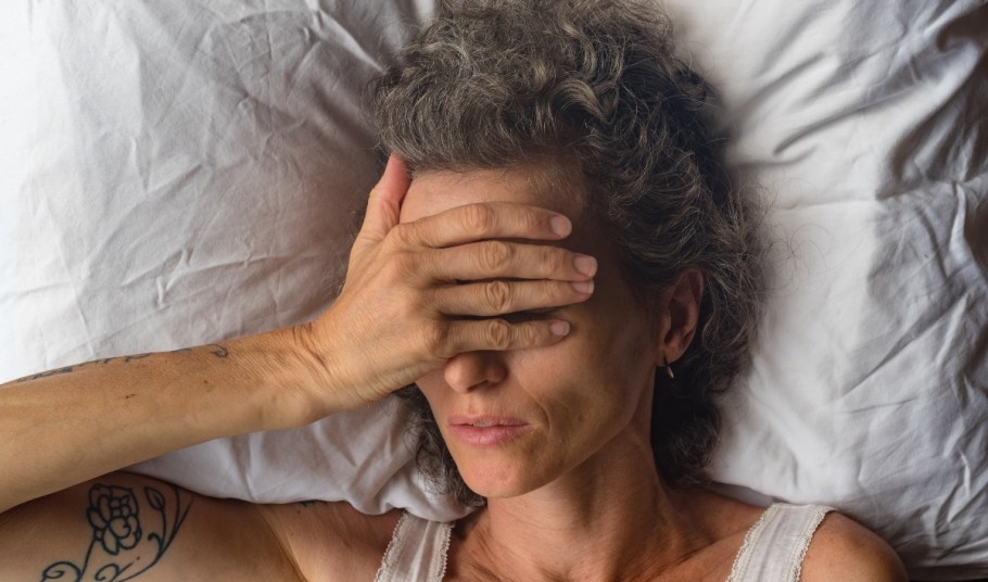 Directly above shot of older woman in bed holding her hand over her face, indicating a headache.