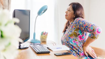 senior woman with a colorful printed shirt sitting at a desk with a computer, pressing hand to back, grimacing from back pain